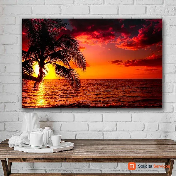 Imagem do acervo com um quadro horizontal de paisagem tropical. Um coqueiro aparece em silhueta diante do mar e de um pôr do sol alaranjado. A peça está acima de uma mesa, em uma parede de tijolos brancos. A textura visível não permite determinar o material estrutural nem o tipo de fixação. Use a cena como referência de decoração; os dados reais da parede precisam ser avaliados.