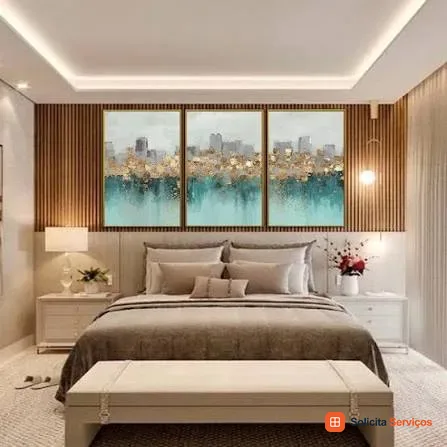 Imagem do acervo do site com três quadros verticais sobre uma cama. As peças mostram uma paisagem urbana e uma área azul-esverdeada na parte inferior. O trio está disposto diante de uma parede com acabamento de madeira. A cabeceira ajuda a observar a proporção e a distribuição do conjunto. Referência de composição; a imagem não informa endereço, autoria da instalação ou medidas.
