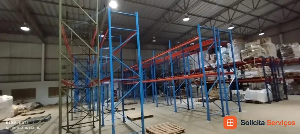 Módulos porta-paletes em um galpão, com montantes azuis e longarinas alaranjadas. Há um andaime ao lado da estrutura, paletes no piso e outras estantes com mercadorias ao fundo.